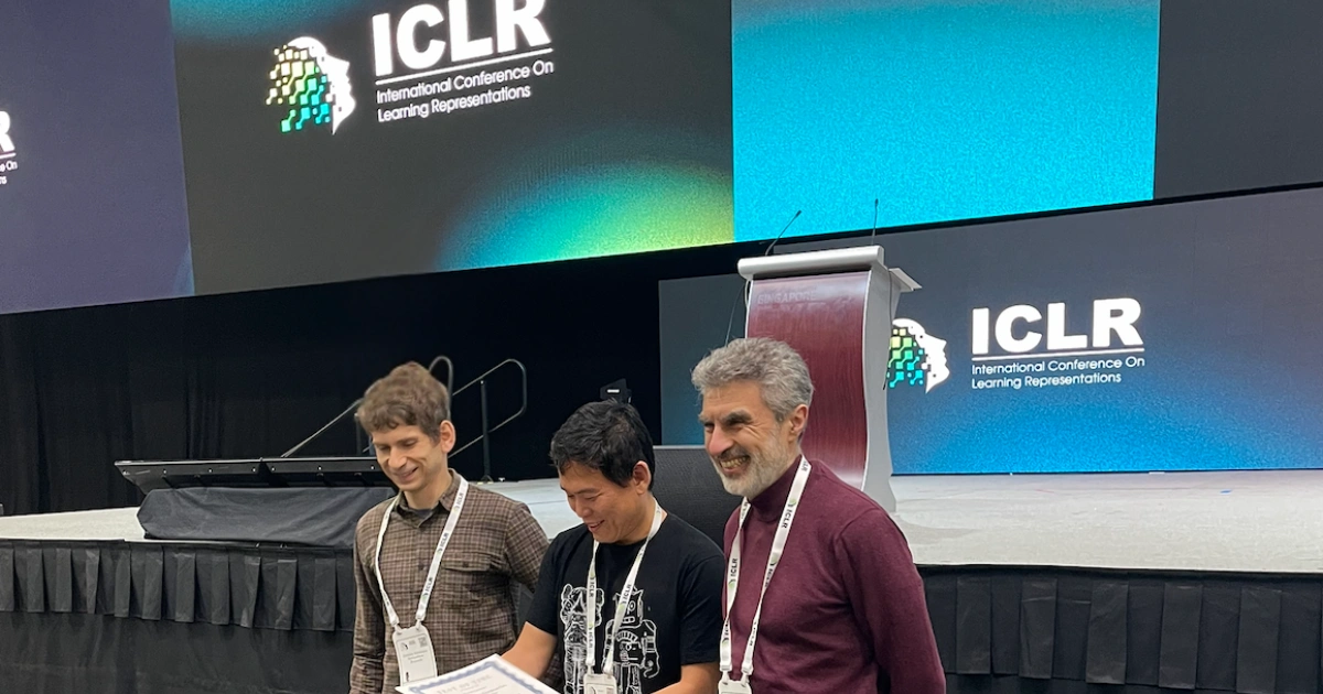 Three people smiling on a stage at a conference with an ICLR banner visible, suggesting a warm and lively event atmosphere.